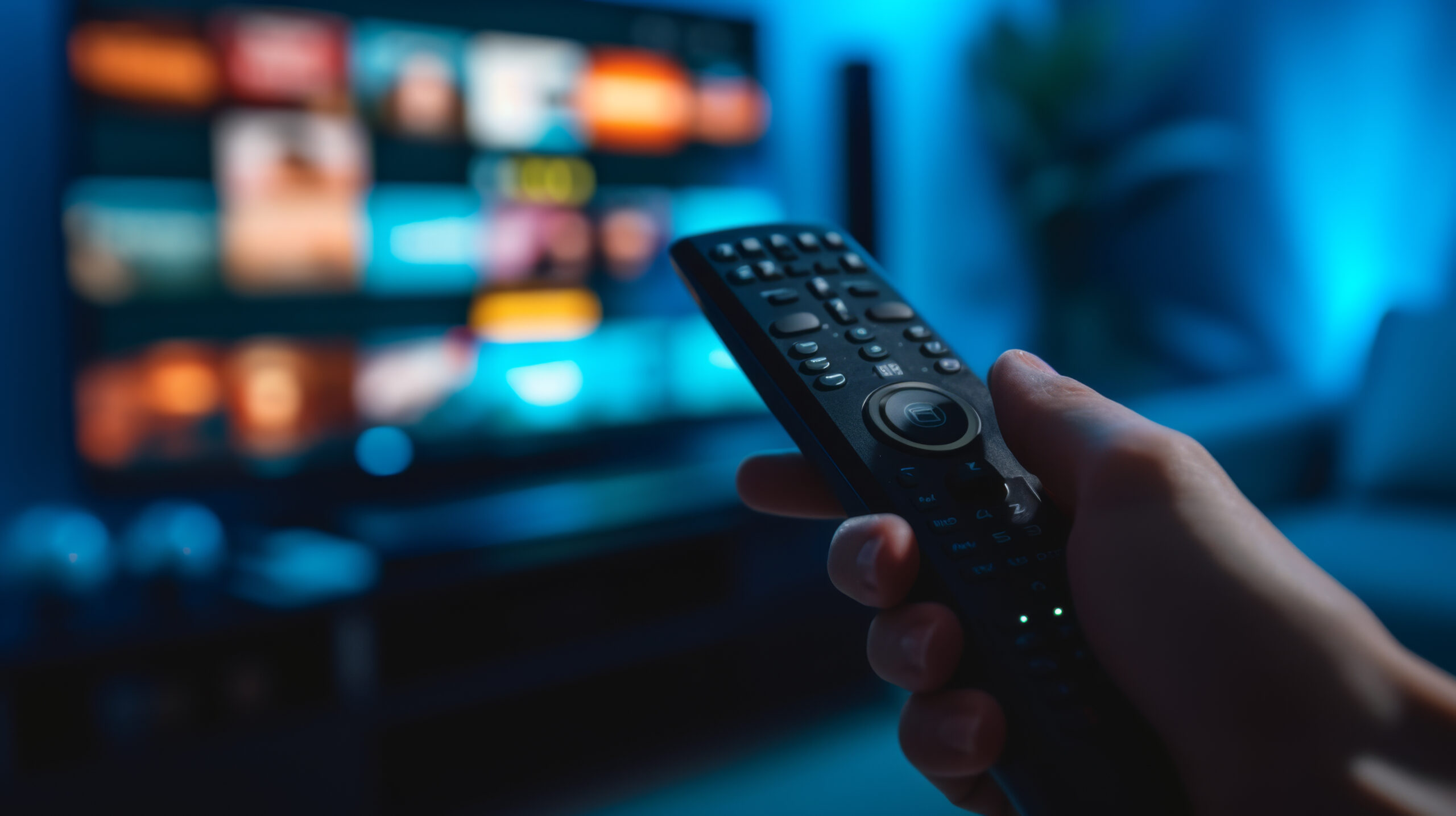 close-up view of a hand holding a television remote control with a blurred television screen in the background, displaying colorful images.